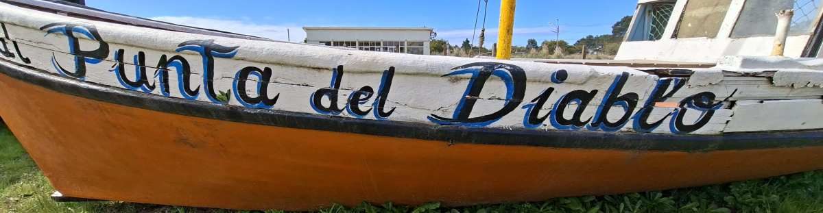 The city name sign is painted on the side of an old boat alongside the road in front of the bus terminal.